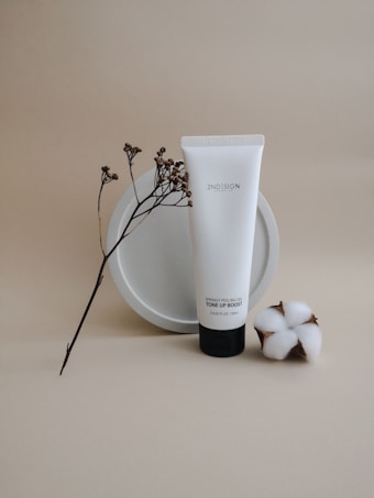 A minimalist still life featuring a white tube of skincare product labeled 'Springy Peeling Gel Tone Up Boost'. It is centered against a neutral beige background, accompanied by a sprig of small dried flowers and a piece of cotton. The arrangement conveys a sense of natural beauty and simplicity.