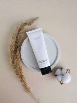 A white tube of skincare product labeled 'Tone Up Boost' lies on a circular ceramic plate with a beige background. Next to it are stalks of dried grasses and a cotton plant.