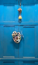 A bright blue wooden door is adorned with a colorful painter's palette and a decorative hanging, which includes a small bell and beads.