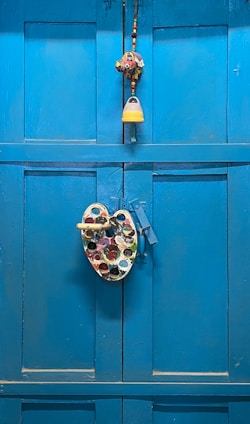 A bright blue wooden door is adorned with a colorful painter's palette and a decorative hanging, which includes a small bell and beads.