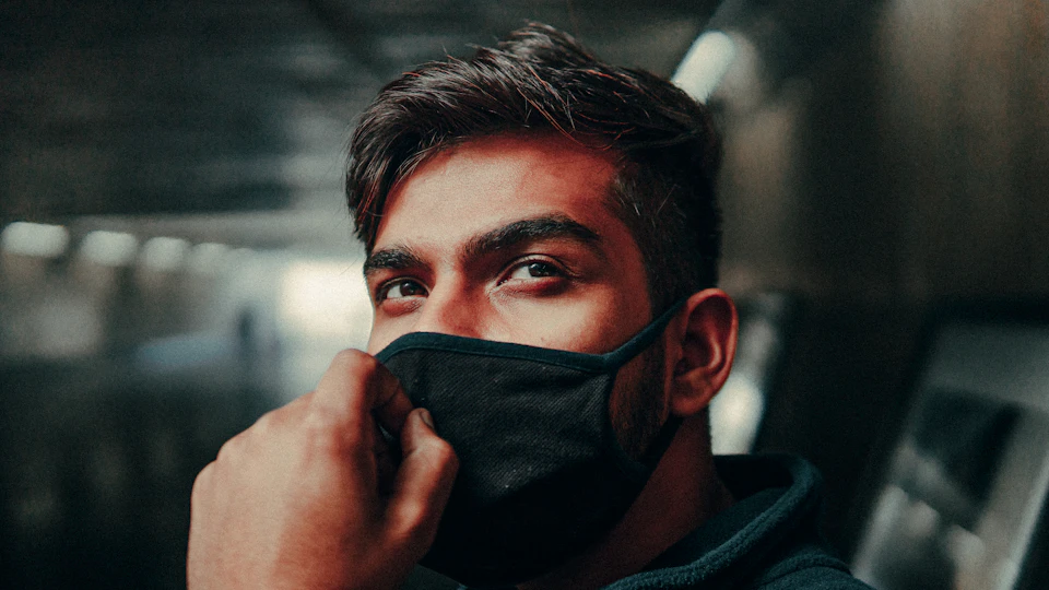 The image shows a man wearing a black face mask, with his eyes looking directly at the camera and his mouth slightly open. The background is dark and out of focus, drawing attention to the subject's face.