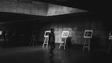 A person wearing dark clothing walks through an art gallery with a series of framed photographs displayed on easels along the wall. The setting is dimly lit, with concrete walls and ceiling, creating a subdued atmosphere.