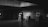 A person wearing dark clothing walks through an art gallery with a series of framed photographs displayed on easels along the wall. The setting is dimly lit, with concrete walls and ceiling, creating a subdued atmosphere.