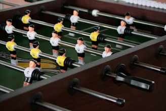 A foosball table with player figures in black and white jerseys, set up in rows on metal rods. The playing surface is green, and the surrounding frame is made of dark wood.