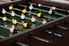 A foosball table with player figures in black and white jerseys, set up in rows on metal rods. The playing surface is green, and the surrounding frame is made of dark wood.