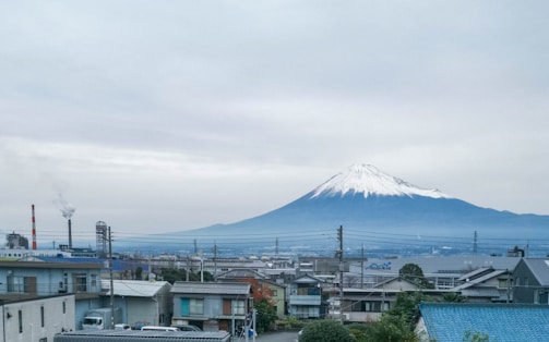 A serene view of Mount Fuji with its snow-capped peak rising above an urban landscape featuring rooftops, electrical wires, and industrial chimneys emitting smoke. The scene is enveloped in a cloudy sky, adding to the tranquil yet industrial contrast.