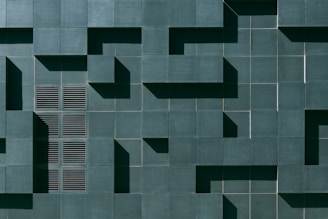 A geometric wall pattern with a series of dark green tiles, some of which protrude to create a three-dimensional effect. The design includes a section with vent-like slats, adding texture and variation to the uniformity.