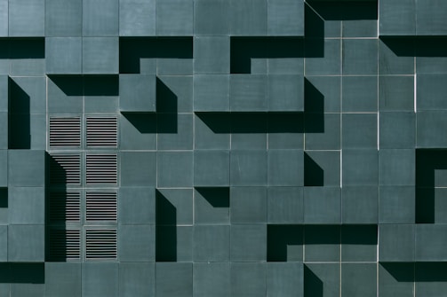 A geometric wall pattern with a series of dark green tiles, some of which protrude to create a three-dimensional effect. The design includes a section with vent-like slats, adding texture and variation to the uniformity.