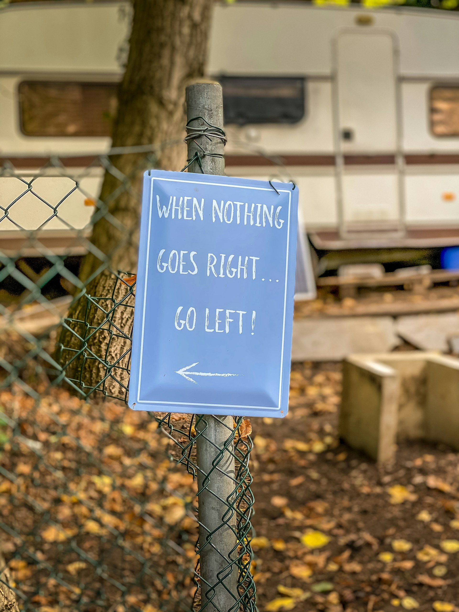 A humorous sign attached to a chain-link fence reads 'When nothing goes right... go left!' with an arrow pointing left. The background features a parked trailer or camper with trees and fallen leaves scattered on the ground, hinting at an outdoor or camping setting.