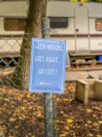 A humorous sign attached to a chain-link fence reads 'When nothing goes right... go left!' with an arrow pointing left. The background features a parked trailer or camper with trees and fallen leaves scattered on the ground, hinting at an outdoor or camping setting.