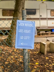 A humorous sign attached to a chain-link fence reads 'When nothing goes right... go left!' with an arrow pointing left. The background features a parked trailer or camper with trees and fallen leaves scattered on the ground, hinting at an outdoor or camping setting.