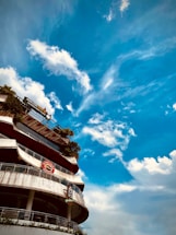 A multi-story building with signage is seen against the backdrop of a bright blue sky with scattered clouds. The building has curved balconies with glass railings and some greenery is present on the upper floors. Notable signage includes 'AVALON BE...' and 'Highla...'.