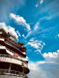 A multi-story building with signage is seen against the backdrop of a bright blue sky with scattered clouds. The building has curved balconies with glass railings and some greenery is present on the upper floors. Notable signage includes 'AVALON BE...' and 'Highla...'.