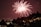 Bright red fireworks burst in the night sky over a cityscape with silhouetted trees and buildings. Street lights and illuminated windows add a warm glow to the scene, enhancing the festive and celebratory atmosphere.