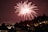 Bright red fireworks burst in the night sky over a cityscape with silhouetted trees and buildings. Street lights and illuminated windows add a warm glow to the scene, enhancing the festive and celebratory atmosphere.