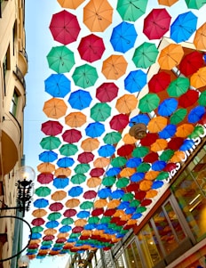 A large number of colorful umbrellas are suspended overhead, creating a canopy effect over a narrow street. The umbrellas are in various colors, including red, blue, green, and orange. The street has a classic street lamp and buildings on both sides, with one building displaying a partially visible sign in the background.