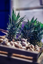 A wooden crate filled with fresh pineapples and ginger roots, illuminated by soft lighting that accentuates the rich green of the pineapple leaves and the earthy tones of the ginger. The background is slightly blurred, emphasizing the produce.