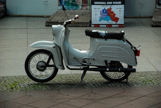A vintage white scooter is parked on a cobblestone and tile pavement. It appears well-used with a slightly worn seat. In the background, a historical map poster titled 'Original DDR Visum' is visible, indicating a setting related to East Germany.