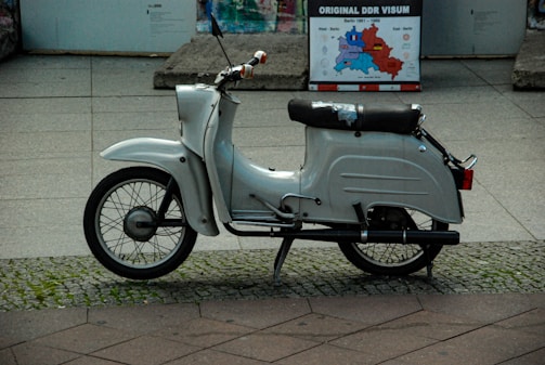 A vintage white scooter is parked on a cobblestone and tile pavement. It appears well-used with a slightly worn seat. In the background, a historical map poster titled 'Original DDR Visum' is visible, indicating a setting related to East Germany.