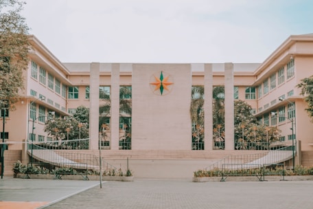 The image features a large, symmetrical building with a central emblem resembling a compass rose. The facade is divided by tall columns, and the structure is flanked by two wings featuring rows of windows with white frames. The courtyard in front is spacious and includes symmetrical, landscaped greenery.