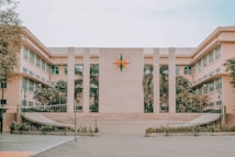 The image features a large, symmetrical building with a central emblem resembling a compass rose. The facade is divided by tall columns, and the structure is flanked by two wings featuring rows of windows with white frames. The courtyard in front is spacious and includes symmetrical, landscaped greenery.