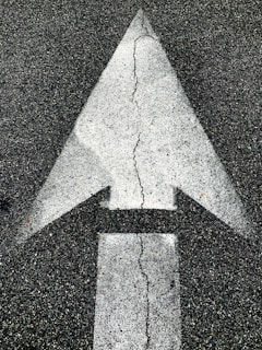 A large white directional arrow painted on a rough, dark asphalt surface with visible cracks running through the arrow.