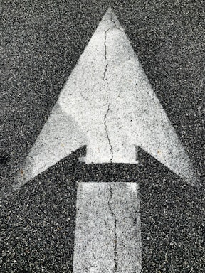A large white directional arrow painted on a rough, dark asphalt surface with visible cracks running through the arrow.