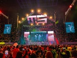 A vibrant concert scene with a large stage featuring colorful lighting and video displays. The stage is decorated with bright lights and the audience is packed with people capturing the event on their phones. Confetti is falling throughout the arena adding to the festive atmosphere.