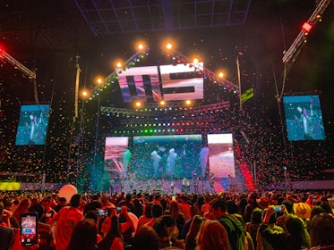 A vibrant concert scene with a large stage featuring colorful lighting and video displays. The stage is decorated with bright lights and the audience is packed with people capturing the event on their phones. Confetti is falling throughout the arena adding to the festive atmosphere.