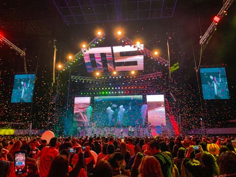 A vibrant concert scene with a large stage featuring colorful lighting and video displays. The stage is decorated with bright lights and the audience is packed with people capturing the event on their phones. Confetti is falling throughout the arena adding to the festive atmosphere.