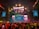 A vibrant concert scene with a large stage featuring colorful lighting and video displays. The stage is decorated with bright lights and the audience is packed with people capturing the event on their phones. Confetti is falling throughout the arena adding to the festive atmosphere.