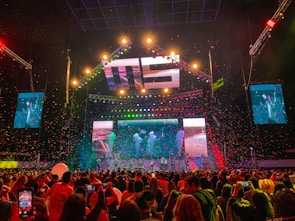 A vibrant concert scene with a large stage featuring colorful lighting and video displays. The stage is decorated with bright lights and the audience is packed with people capturing the event on their phones. Confetti is falling throughout the arena adding to the festive atmosphere.