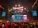 A vibrant concert scene with a large stage featuring colorful lighting and video displays. The stage is decorated with bright lights and the audience is packed with people capturing the event on their phones. Confetti is falling throughout the arena adding to the festive atmosphere.