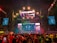 A vibrant concert scene with a large stage featuring colorful lighting and video displays. The stage is decorated with bright lights and the audience is packed with people capturing the event on their phones. Confetti is falling throughout the arena adding to the festive atmosphere.