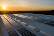 A series of solar panels are installed on a large industrial rooftop under a clear sky at sunset. The sunlight casts a warm glow across the panels and the surrounding area, highlighting the geometric patterns formed by the panels.