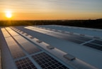 A series of solar panels are installed on a large industrial rooftop under a clear sky at sunset. The sunlight casts a warm glow across the panels and the surrounding area, highlighting the geometric patterns formed by the panels.