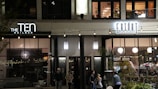 A street view of a modern building façade at night, featuring two businesses: a nail bar on the left and a market on the right, with people walking by. The businesses have large windows and are illuminated with signage and string lights. The upper floor has windows with visible interior and plants.
