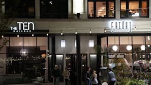 A street view of a modern building façade at night, featuring two businesses: a nail bar on the left and a market on the right, with people walking by. The businesses have large windows and are illuminated with signage and string lights. The upper floor has windows with visible interior and plants.