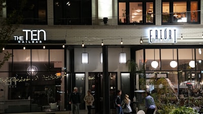 A street view of a modern building fa&ccedil;ade at night, featuring two businesses: a nail bar on the left and a market on the right, with people walking by. The businesses have large windows and are illuminated with signage and string lights. The upper floor has windows with visible interior and plants.