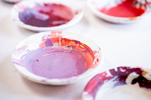 Several small dishes are filled with various shades of paint, including purple, red, and other vibrant colors. The dishes are placed on a white surface, and the paint appears to be mixed or used, giving an artistic and creative atmosphere.