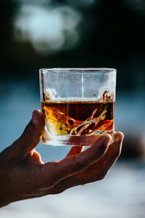 Close-up of a hand holding a whiskey glass with amber liquid, set against a blurred warm bar background.