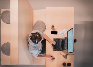 A person wearing a gray hoodie and headphones is sitting at a desk, working on a laptop with an external monitor. Nearby are a smartphone, an open notebook, a face mask, and a beverage bottle. The workspace is organized on a light wooden surface with additional seating to the side.