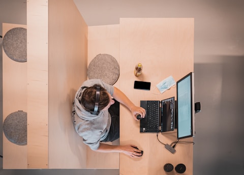 A person wearing a gray hoodie and headphones is sitting at a desk, working on a laptop with an external monitor. Nearby are a smartphone, an open notebook, a face mask, and a beverage bottle. The workspace is organized on a light wooden surface with additional seating to the side.