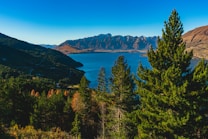 A breathtaking landscape of a lake surrounded by mountains with lush green trees in the foreground. The mountains are rugged and majestic with a mix of shadows and sunlight accentuating their texture. The deep blue of the sky complements the clear lake, creating a peaceful and serene atmosphere.