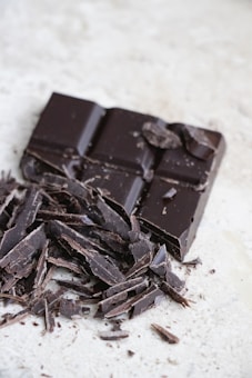 A block of dark chocolate is partially broken with several shards scattered around on a light-colored surface.