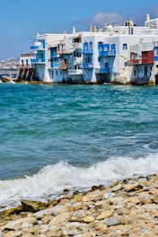 Seaside buildings with white walls and colorful balconies, next to a clear blue ocean with waves breaking on a rocky shoreline.