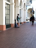 A street musician stands next to a beige building with arched windows, playing a blue guitar. A backpack and other belongings rest on the ground beside him. Pedestrians walk nearby on the brick sidewalk, passing under a shop sign with 'Brandy Melville' written on it. The scene takes place in an urban environment with cars and a traffic light visible in the background.