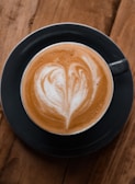 A cup of coffee with intricate latte art on top, consisting of a heart-shaped pattern made with steamed milk. The coffee is in a dark-colored mug placed on a wooden surface.
