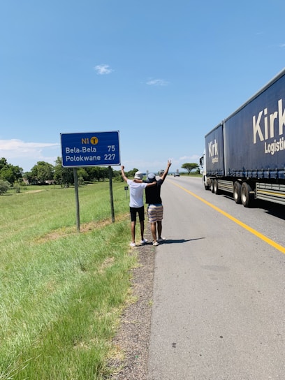 Two individuals stand on the side of a highway, facing away from the camera with arms raised. They are next to a road sign indicating directions and distances to Bela-Bela and Polokwane. A large truck labeled 'Kirk Logistics' is moving on the road. The scene is set in a rural area with green grass and trees under a clear blue sky.