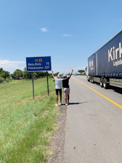 Two individuals stand on the side of a highway, facing away from the camera with arms raised. They are next to a road sign indicating directions and distances to Bela-Bela and Polokwane. A large truck labeled 'Kirk Logistics' is moving on the road. The scene is set in a rural area with green grass and trees under a clear blue sky.